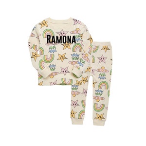 May include: A cream-colored pajama set with a long-sleeved top and pants. The top has a colorful print of stars, rainbows, and UFOs. The pants have the same print. The top has the name "RAMONA" printed on the front.