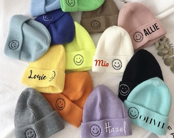May include: A collection of 12 colorful knitted beanies with a smiley face design and personalized names, including Lorie, Mia, Scout, Allie, Hazel, Oliver.