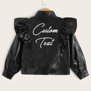 以下が含まれることがあります： 肩にフリルが付いた黒いフェイクレザーのジャケット。ジャケットはジッパーで閉まり、白い刺繍で「Custom Text」と書いてあります。