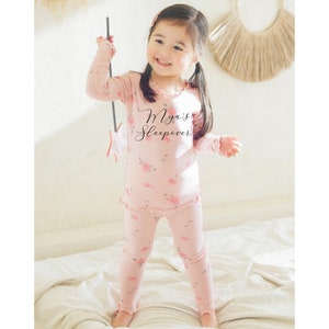 May include: A pink long-sleeved pajama top and matching pants with a floral print. The top has a ruffled neckline and the text "Mya's Sleepover" printed on the front.