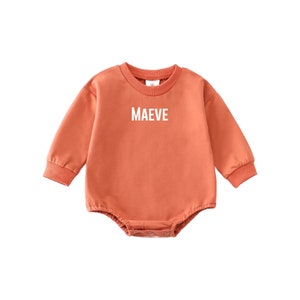 May include: A long-sleeved, orange baby onesie with the name "MAEVE" printed in white on the front.