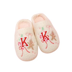 May include: A pair of cream-colored, plush slippers. Each slipper features a red letter "K", a gingerbread man, and festive red and pink accents. The slippers have a soft, pink interior.