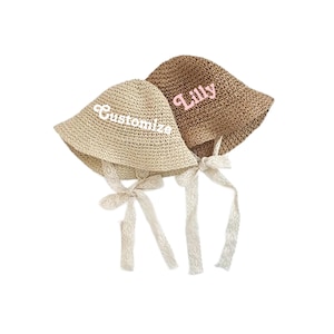 May include: Two straw sun hats with white lace ties. One hat is beige with the word "Customize" embroidered in white. The other hat is brown with the word "Lilly" embroidered in pink.