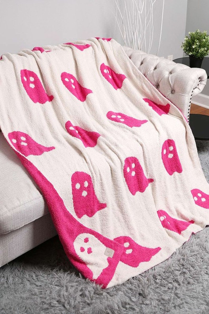 K&ouml;nnte beinhalten: Eine wei&szlig;e Decke mit einem Muster aus rosa Geistern. Die Decke ist gefaltet und &uuml;ber ein wei&szlig;es Sofa gelegt.