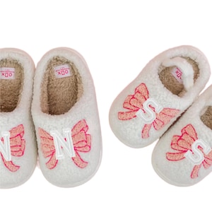 May include: White plush slippers with pink bow accents and embroidered letters. The slippers have a soft, fuzzy interior and are designed for indoor wear. The letters "N" and "S" are visible on the slippers.