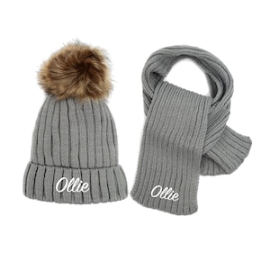 May include: Gray knitted hat with a brown faux fur pom pom and matching gray scarf. Both items have the name "Ollie" embroidered in white.