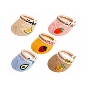 May include: Five straw visors in beige, pink, yellow, light blue, and tan. Each visor features a striped headband with a personalized name and a colorful embroidered fruit patch, including a lime, carrot, strawberry, avocado, and pineapple.