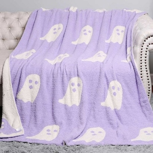K&ouml;nnte beinhalten: Eine weiche, lilafarbene Kuscheldecke mit einem wei&szlig;en Geistermuster. Die Decke ist gefaltet und &uuml;ber ein wei&szlig;es Sofa gelegt.
