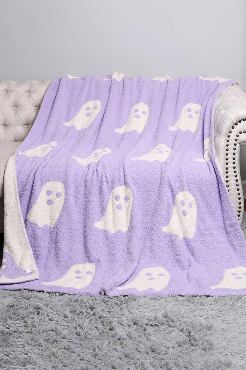 K&ouml;nnte beinhalten: Eine weiche, lilafarbene Kuscheldecke mit einem wei&szlig;en Geistermuster. Die Decke ist gefaltet und &uuml;ber ein wei&szlig;es Sofa gelegt.