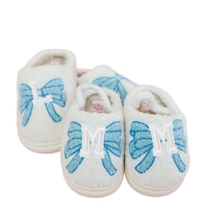 May include: White plush slippers with light blue bow accents and embroidered letters. One slipper has the letter "L", and two have the letter "M". The slippers are designed for indoor use and have a soft, comfortable appearance.
