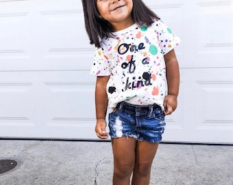 May include: A young girl wearing a white t-shirt with colorful paint splatters and the text "One of a kind". She is also wearing denim shorts and white sneakers. She has a large orange bow in her hair.