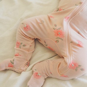 May include: A pink baby sleeper with a floral print. The sleeper has a zipper down the front and footie feet.
