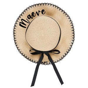 May include: A straw sun hat with a black ribbon and trim. The hat features the name "Maeve" in black script across the brim. The hat has a wide brim and a round crown, perfect for sun protection.