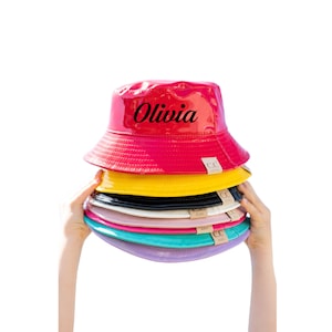 May include: A stack of colorful, shiny bucket hats. The top hat is red and has the name "Olivia" written on it in black.