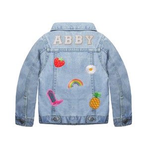 Op de afbeelding: Een lichtblauw denim jack met de naam "ABBY" geborduurd op de rug in witte letters. Het jack heeft verschillende kleurrijke patches genaaid op de rug, waaronder een aardbei, een madeliefje, een regenboog, een ananas en een roze cowboylaars.