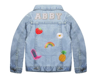 Könnte beinhalten: Eine hellblaue Jeansjacke mit dem Namen "ABBY" in weißen Buchstaben auf der Rückseite gestickt. Die Jacke hat mehrere bunte Patches auf der Rückseite aufgenäht, darunter eine Erdbeere, eine Gänseblümchen, ein Regenbogen, eine Ananas und ein rosa Cowboy-Stiefel.