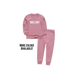 May include: A pink long-sleeved shirt and matching pants pajamas set for children. The shirt has the name "Mallory" printed in white on the front. The text "More Colors Available!" is printed below the image.