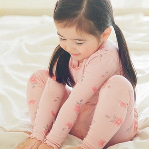 May include: A young girl wearing a pink long-sleeved pajamas with a floral print. The pajamas have ruffles at the wrists and ankles.