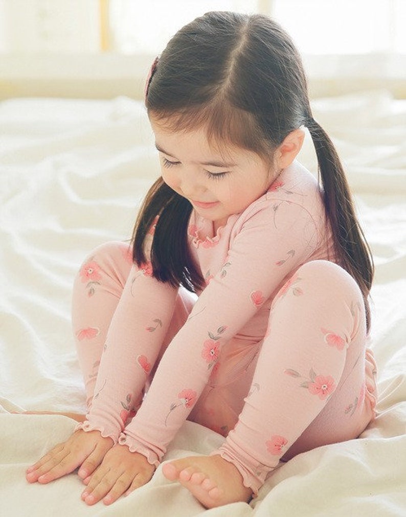 May include: A young girl wearing a pink long-sleeved pajamas with a floral print. The pajamas have ruffles at the wrists and ankles.