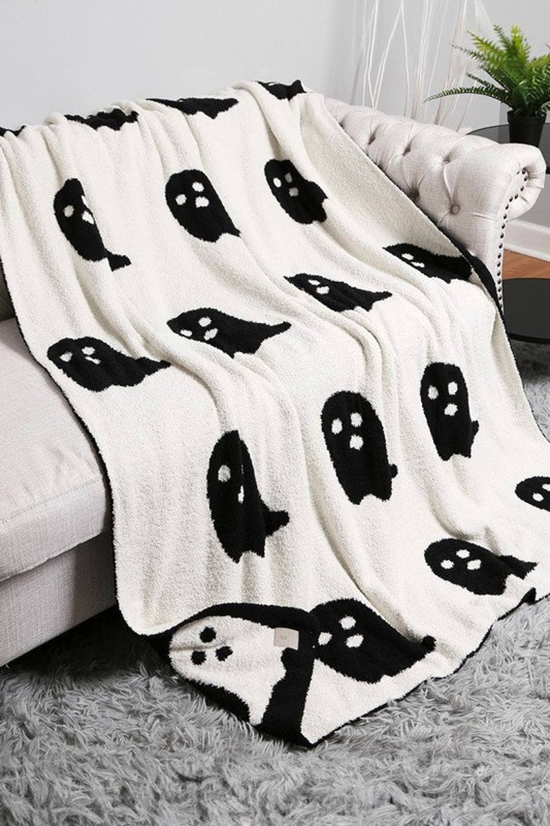 K&ouml;nnte beinhalten: Eine wei&szlig;e Kuscheldecke mit einem schwarzen Geistermuster. Die Decke ist weich und flauschig und perfekt zum Kuscheln auf der Couch.