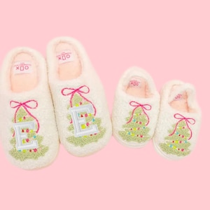 May include: Four pairs of white, fluffy Christmas slippers. Two adult-sized slippers feature an embroidered green Christmas tree with colorful lights and a pink bow. The other two are child-sized slippers with a Christmas tree design.
