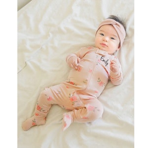 May include: A pink baby onesie with a floral print and the name "Emily" embroidered on the front. The onesie has long sleeves and feet.