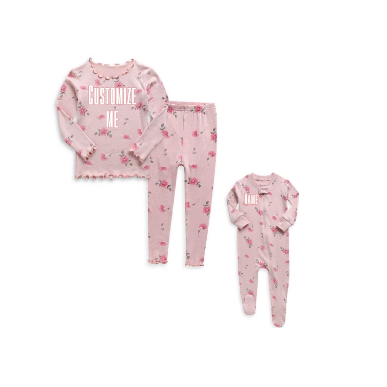May include: Pink pajamas with a floral print. The top has the text "CUSTOMIZE ME" on it. The bottom is a pair of pants. The second pajama set is a pink one-piece with the text "NAME" on it.