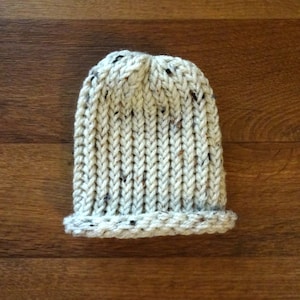 May include: A white knitted beanie with a flecked texture. The beanie has a ribbed pattern and a folded brim.