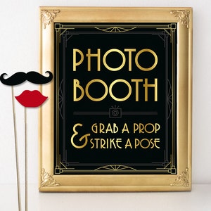 May include: A framed photo booth sign with gold lettering on a black background. The sign reads "PHOTO BOOTH" and "GRAB A PROP & STRIKE A POSE". A black mustache and red lips on sticks are in the foreground.