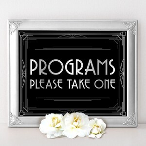 Peut inclure: Une pancarte encadrée argent avec un fond noir et un design art déco blanc. La pancarte indique "Programmes Veuillez prendre un".
