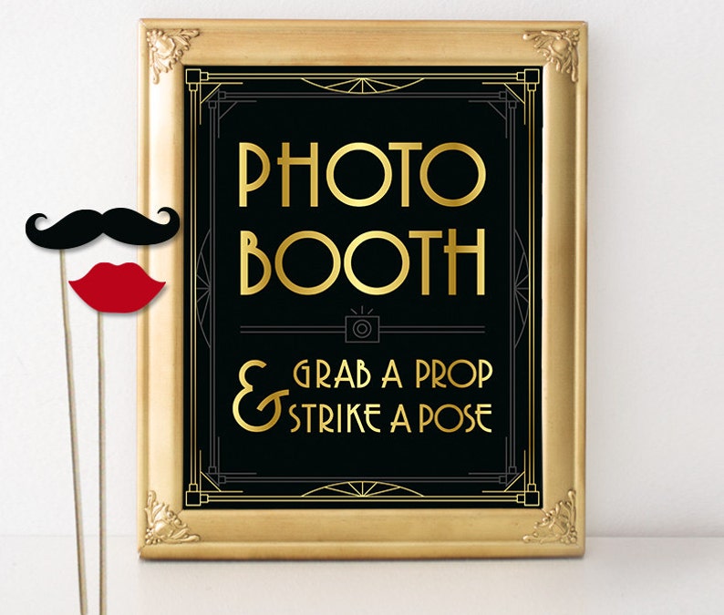 May include: A framed photo booth sign with gold lettering on a black background. The sign reads "PHOTO BOOTH" and "GRAB A PROP & STRIKE A POSE". A black mustache and red lips on sticks are in the foreground.