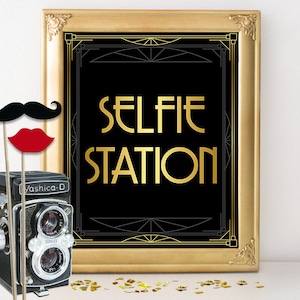 May include: A gold framed sign with black background and gold lettering that reads "SELFIE STATION". The sign is sitting on a white surface with gold confetti scattered around it. There is a vintage camera and a mustache and red lips on sticks in the foreground.