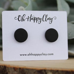 May include: A pair of matte black circular stud earrings displayed on a white rectangular card. The card features the black text Oh Happy Clay at the top and the website address www.ohhappyclay.com printed in black at the bottom center.