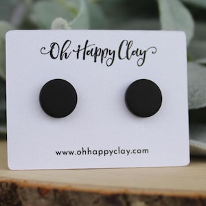 May include: A pair of matte black circular stud earrings displayed on a white rectangular card. The card features the black text Oh Happy Clay at the top and the website address www.ohhappyclay.com printed in black at the bottom center.