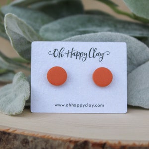 May include: A pair of round, orange stud earrings displayed on a white card with the text "Oh Happy Clay" and the website address. The earrings are set against a backdrop of green foliage and a wooden surface.