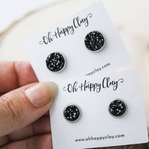 Puede incluir: Dos pares de pendientes de botón druzy negros sobre tarjetas blancas. Los pendientes tienen un bisel plateado. Las tarjetas tienen el texto "Oh Happy Clay" y "ohhappyclay.com" impreso. Los pendientes son sostenidos por una mano.