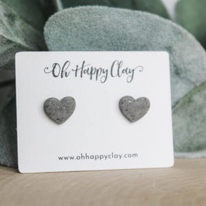 May include: A pair of gray heart-shaped stud earrings on a white card with the text "Oh Happy Clay" and the website address. The earrings are made of a speckled material and are displayed against a backdrop of green foliage.
