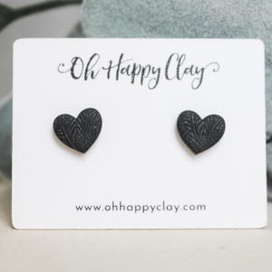 May include: A pair of black heart-shaped stud earrings with a textured surface on a white display card. The card features the text Oh Happy Clay at the top and the website www.ohhappyclay.com printed in black at the bottom center.