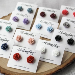May include: A collection of rose-shaped stud earrings in various colors, including blue, pink, red, and white, displayed on white cards. The cards are arranged on a wooden surface, with the text "Oh Happy Clay" and a website address visible.