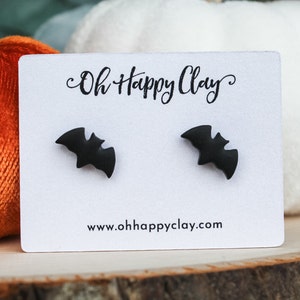 Puede incluir: Un par de pendientes de botón en forma de murciélago de color negro mate sujetos a una tarjeta de presentación blanca. La tarjeta presenta el texto Oh Happy Clay en una fuente cursiva negra en la parte superior y la web www.ohhappyclay.com en negro.