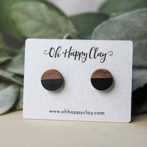 May include: A pair of wooden earrings with a black and brown wood design. The earrings are displayed on a white card with the text "Oh Happy Clay" and "www.ohhappyclay.com".