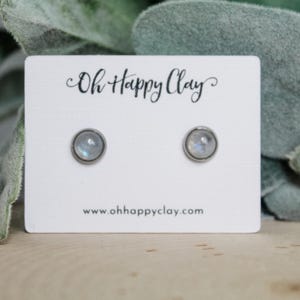 May include: A pair of round stud earrings with a silver-colored setting and a light blue and white stone. The earrings are displayed on a white card with the text "Oh Happy Clay" and the website address www.ohhappyclay.com.