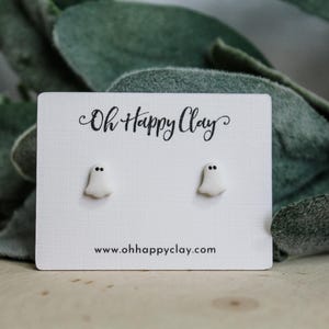 Puede incluir: Un par de pendientes de botón con forma de fantasma blanco y ojos de punto negro sobre una tarjeta expositora blanca. La tarjeta muestra el texto negro Oh Happy Clay arriba y el sitio web www.ohhappyclay.com abajo. Las formas son lisas.