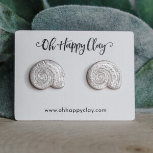 May include: A pair of silver-colored seashell-shaped stud earrings on a white card with the text "Oh Happy Clay" and the website address www.ohhappyclay.com. The earrings have a spiral design. The background features green foliage.