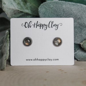 May include: A pair of stud earrings with a silver-colored setting and a round, iridescent stone. The earrings are displayed on a white card with the text "Oh Happy Clay" and the website address. The background includes green foliage.