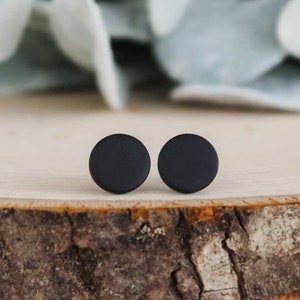 Puede incluir: Un par de pendientes de bot&oacute;n circulares en color negro mate. Cada pendiente presenta un dise&ntilde;o sencillo, plano y redondo con una superficie lisa y no reflectante. Los pendientes est&aacute;n colocados uno al lado del otro, mostrando su forma.
