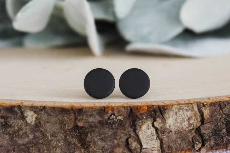 Puede incluir: Un par de pendientes de bot&oacute;n circulares en color negro mate. Cada pendiente presenta un dise&ntilde;o sencillo, plano y redondo con una superficie lisa y no reflectante. Los pendientes est&aacute;n colocados uno al lado del otro, mostrando su forma.