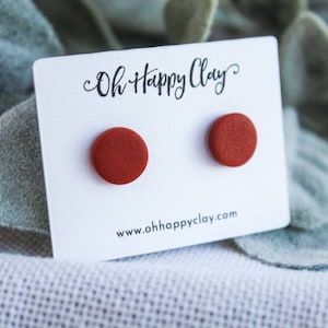 May include: A pair of round, terracotta-colored stud earrings on a white card with the text "Oh Happy Clay" and the website address. The earrings are displayed against a backdrop of soft, green foliage.