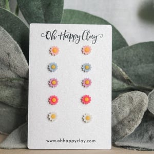 May include: A white card displays a collection of daisy stud earrings in various colors: peach, blue, pink, red, and yellow. The card is printed with "Oh Happy Clay" and the website address. The earrings are set against a backdrop of green foliage.