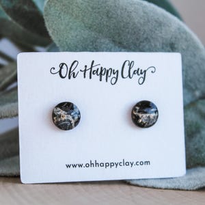 May include: A pair of round stud earrings with a marbled black, white, and gold design. The earrings are displayed on a white card with the text "Oh Happy Clay" and the website address www.ohhappyclay.com.